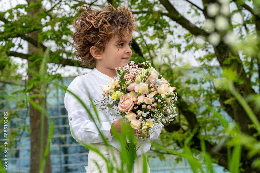 A curly-haired child in a white dress holds a bouquet of flowers while standing outdoors amidst lush greenery. A curly-haired child in a white dress holds a bouquet of flowers while standing outdoors amidst lush greenery.