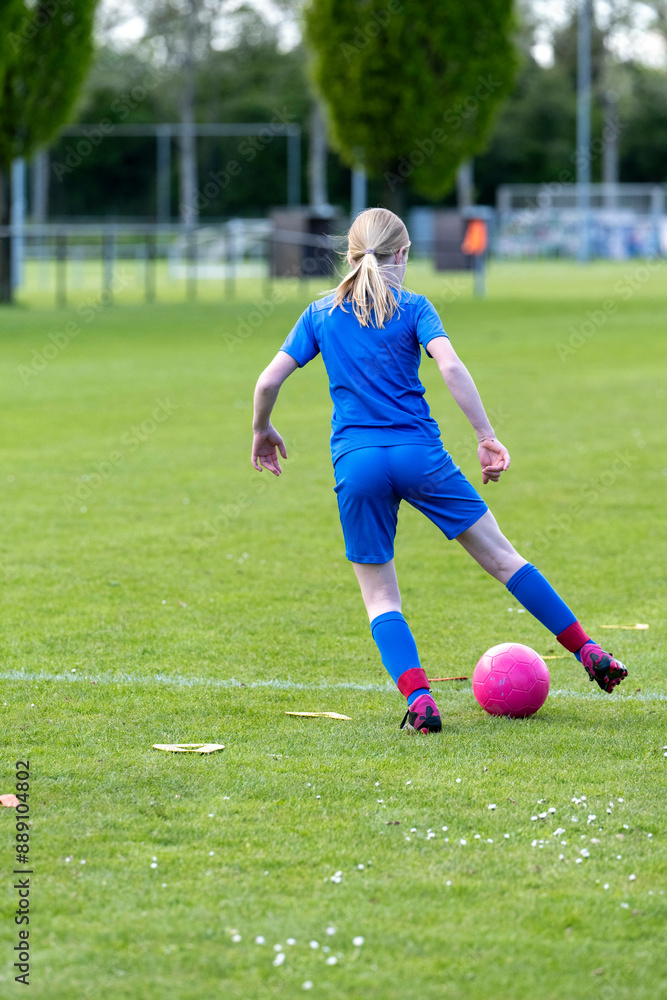 A female soccer player in a blue jersey dribbles a pink soccer ball on a green grass field. A female soccer player in a blue jersey dribbles a pink soccer ball on a green grass field.