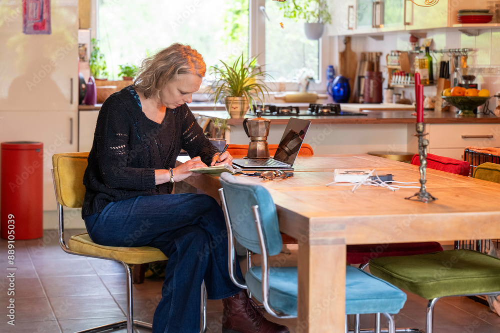Adult woman writing in a notebook at a wooden dining table with kitchen background. Adult woman writing in a notebook at a wooden dining table with kitchen background.