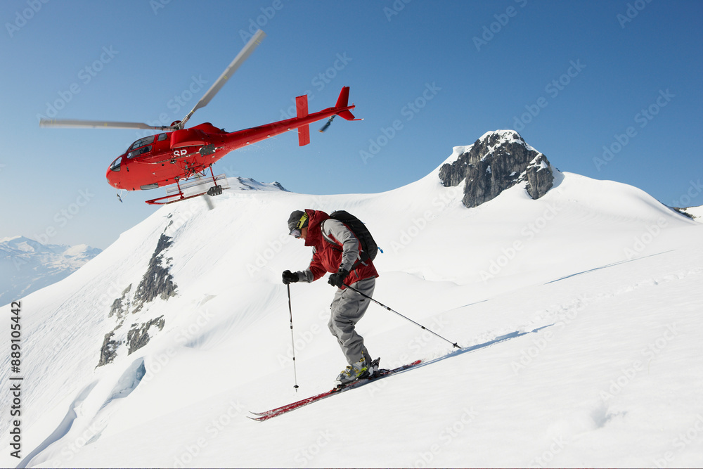 Skier in red descending a pristine mountain slope with a red helicopter in flight in the backdrop. Skier in red descending a pristine mountain slope with a red helicopter in flight in the backdrop.