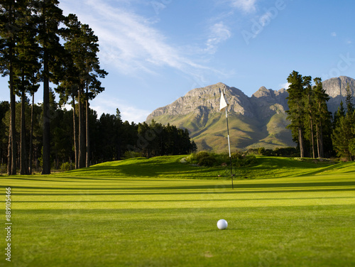 Golf ball on a lush green putting green with flagstick and dramatic mountain background under a clear blue sky. photo