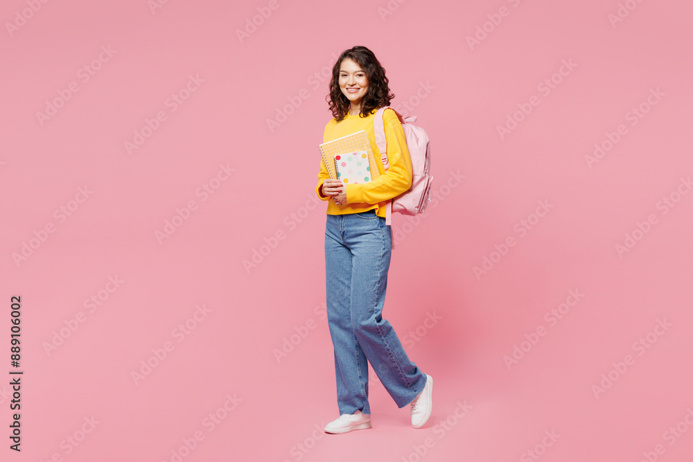 Full body side view young smart teen girl student she wearing yellow casual clothes backpack bag holding books walk go isolated on plain pastel pink background. High school university college concept. Full body side view young smart teen girl student she wearing yellow casual clothes backpack bag holding books walk go isolated on plain pastel pink background. High school university college concept.
