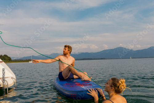 Man sitting on a wakeboard being handed a tow rope by a woman from a boat on a calm lake with mountains in the background.