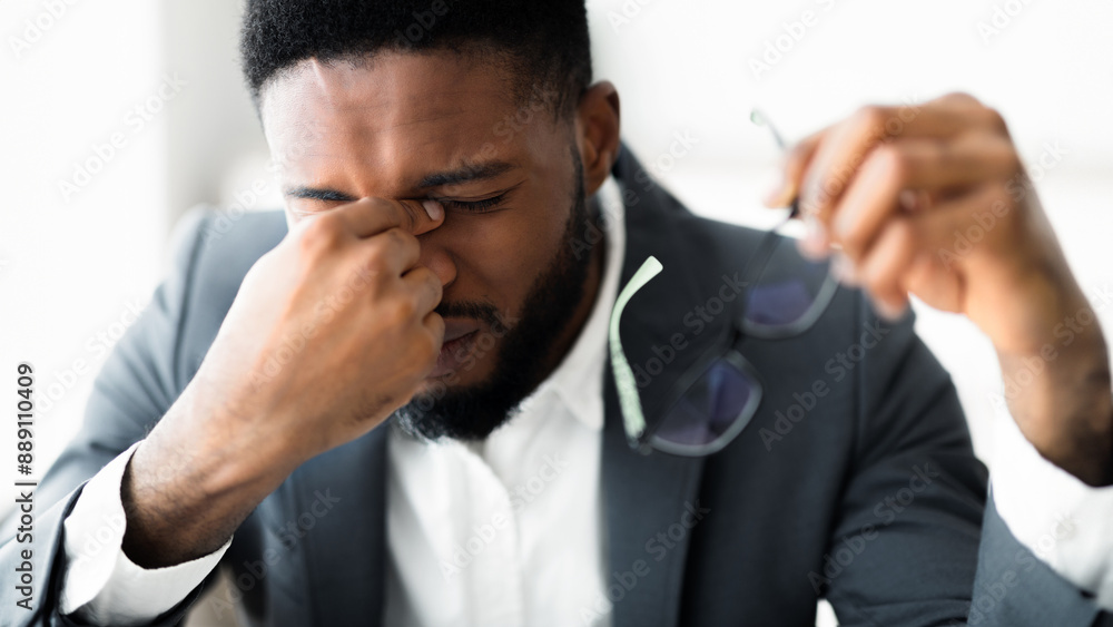 A man in a suit rubs his eyes with his hand. He holds glasses in his other hand. A man in a suit rubs his eyes with his hand. He holds glasses in his other hand.