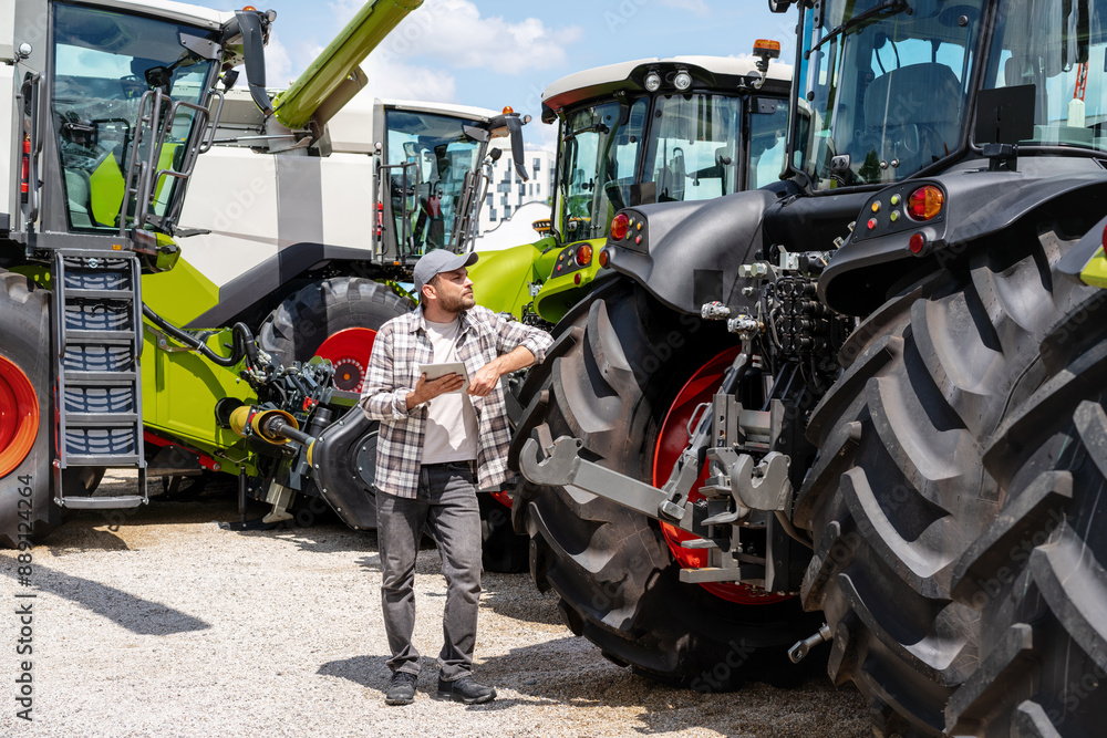 Farm machinery dealer. A man with a digital tablet stands at an agricultural equipment dealer and looks at tractor. Farm machinery dealer. A man with a digital tablet stands at an agricultural equipment dealer and looks at tractor.