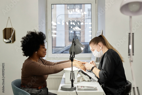 Side view shot of female nail master working on manicure of happy African American client with curly hair. Black woman at nail tech desk getting nails cut, filed, and shaped, copy space