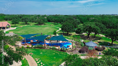 Slash pad with sail shade structure and diverse kids playing water in large modern children recreation center, lush green tree to horizontal line, suburban park, Flower Mound, Texas, aerial view