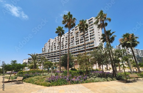 Casa Finance City in Casablanca, Morocco, featuring a modern skyscraper with distinctive architecture. The scene includes a lush garden with purple flowers and palm trees under a clear blue sky