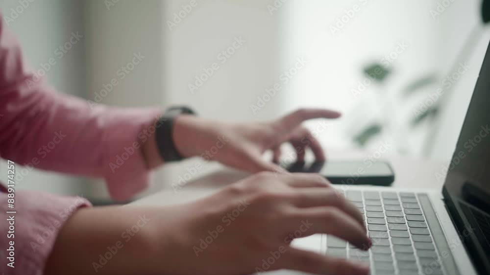 An Asian man in a pink shirt is working on his laptop in an office while taking a phone call. He appears focused, with documents on the desk. The scene depicts a typical busy workday. An Asian man in a pink shirt is working on his laptop in an office while taking a phone call. He appears focused, with documents on the desk. The scene depicts a typical busy workday.