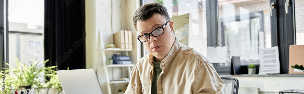 A young man with Down syndrome sits at a desk in an office, working on his laptop. A young man with Down syndrome sits at a desk in an office, working on his laptop.