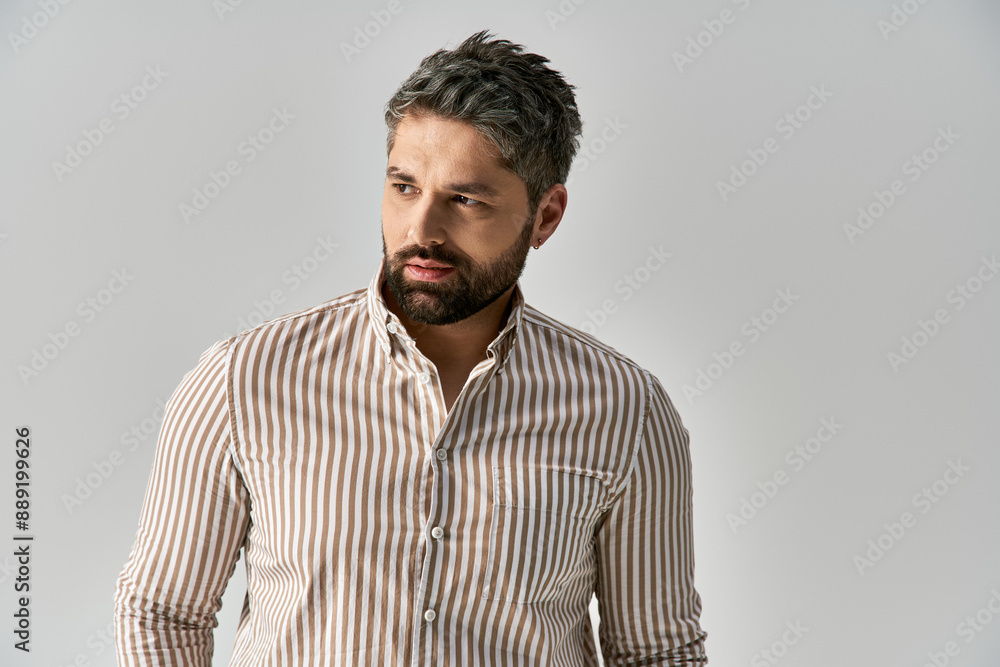 Obraz premium A bearded man in an elegant striped shirt strikes a pose against a grey studio background.