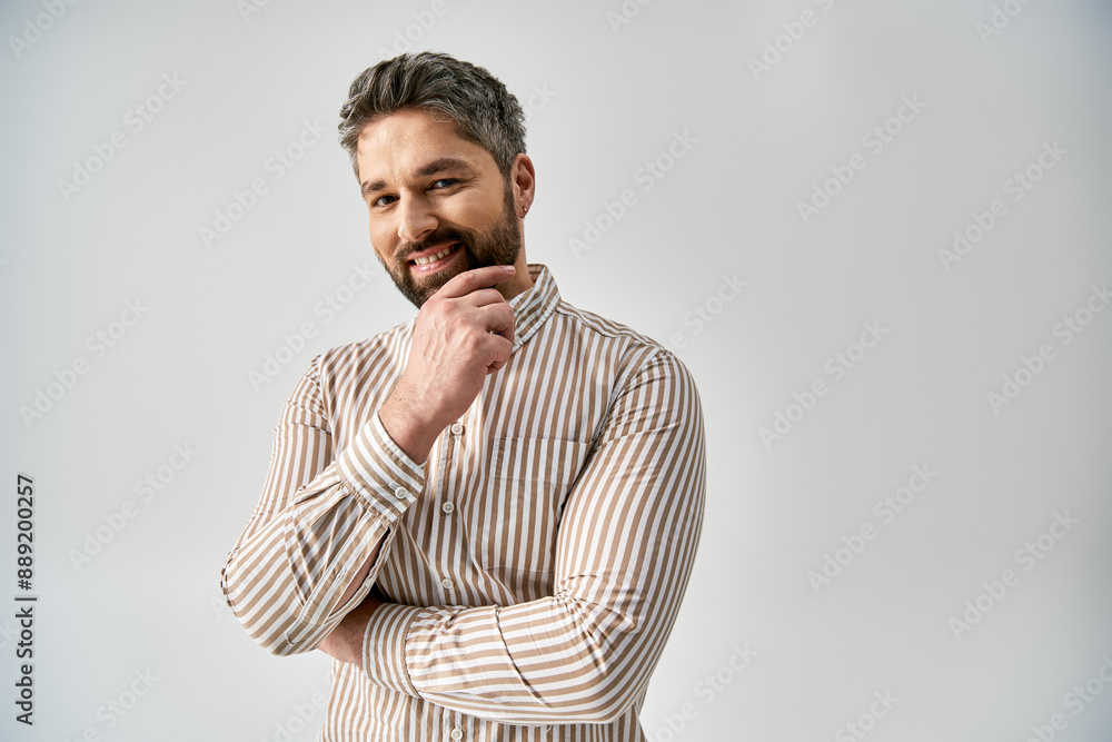 A stylish man with a beard poses in elegant attire against a grey background in a studio setting. A stylish man with a beard poses in elegant attire against a grey background in a studio setting.