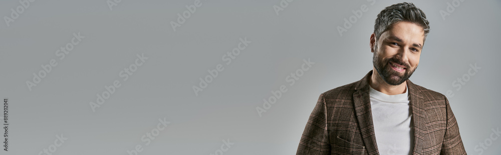 Obraz premium A stylish bearded man in elegant attire stands confidently in front of a sleek gray backdrop in a studio setting.