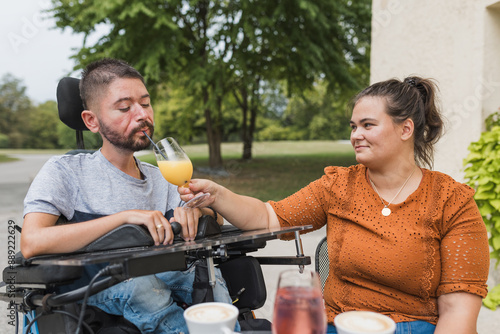 Caring wife helping her husband with a disability drink juice while enjoying sitting in the cafe garden. Love and commitment concepts. photo