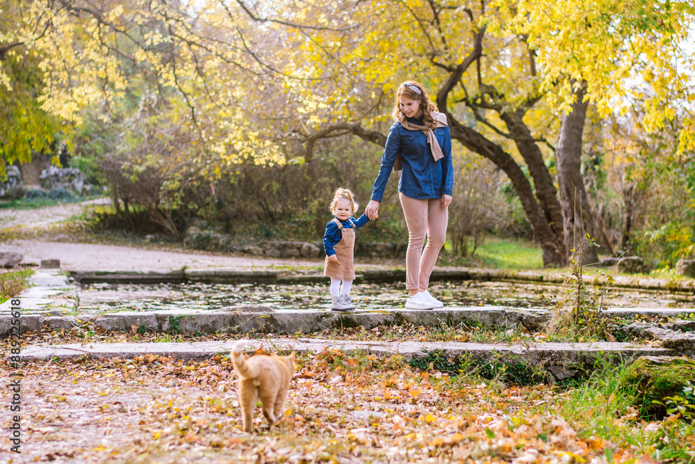 mother and daughter walk hand-in-hand through park autumn leaves. girl wearing brown dress. mother wearing blue shirt and tan pants. time with mother. happy childhood. maternity leave to care child mother and daughter walk hand-in-hand through park autumn leaves. girl wearing brown dress. mother wearing blue shirt and tan pants. time with mother. happy childhood. maternity leave to care child