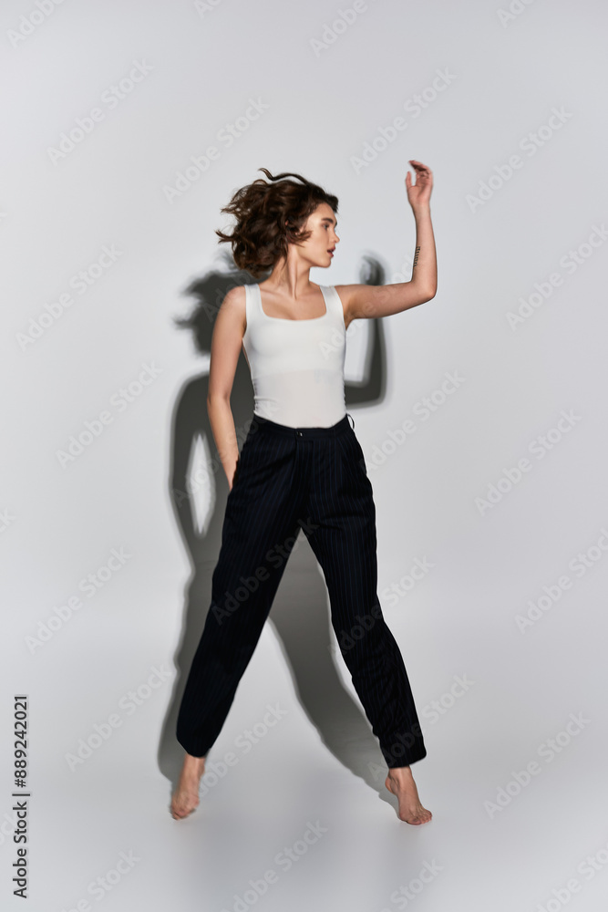A pretty young woman strikes a graceful pose in black pants and white tank top against a grey studio backdrop. A pretty young woman strikes a graceful pose in black pants and white tank top against a grey studio backdrop.