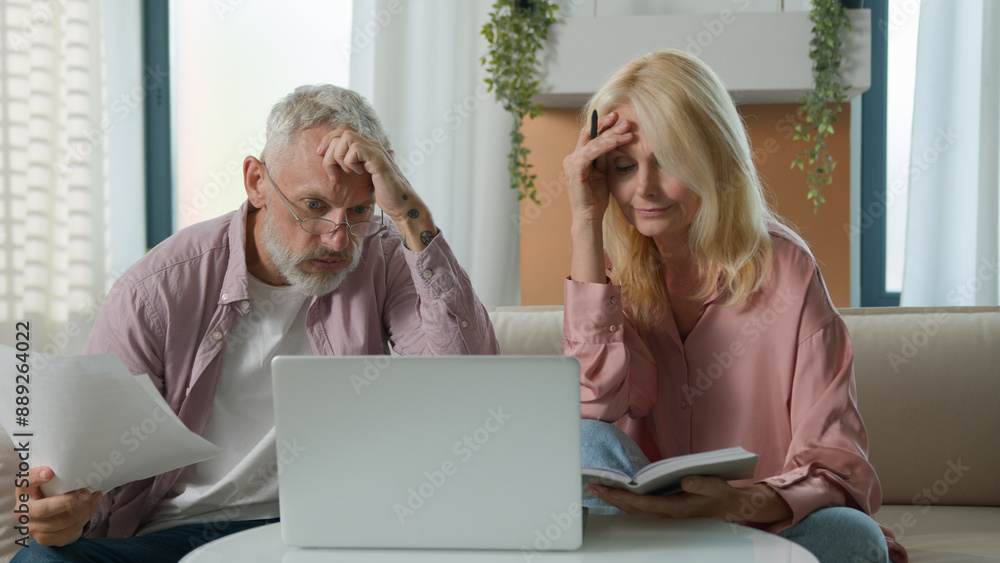 © Yuliia - Angry upset 50s married senior elderly Caucasian couple woman female mature man male businesspeople family bankruptcy business documents paper laptop speak bill money debt problem scam home bad result