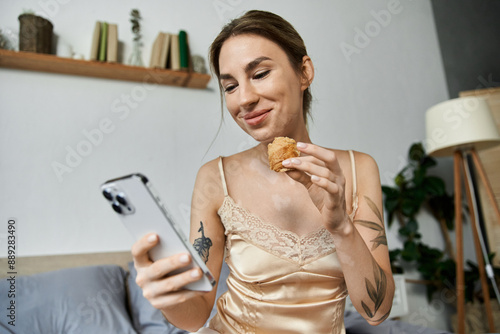 A young woman with vitiligo smiles as she eats a pastry while using her phone in her cozy bedroom. photo