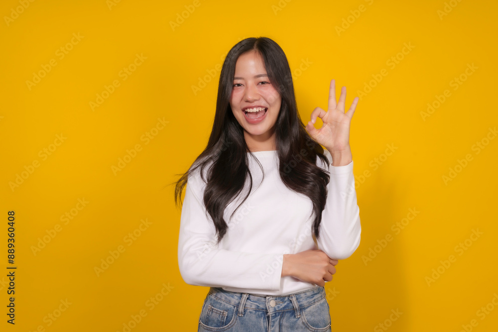 Young woman happily shows approval with an ok sign against a yellow backdrop, radiating positivity and satisfaction Young woman happily shows approval with an ok sign against a yellow backdrop, radiating positivity and satisfaction