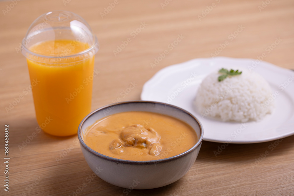 Delicious bowl of creamy butter chicken served with a mound of steamed white rice garnished with cilantro on a white plate, accompanied by a refreshing orange juice in a plastic cup. Delicious bowl of creamy butter chicken served with a mound of steamed white rice garnished with cilantro on a white plate, accompanied by a refreshing orange juice in a plastic cup.