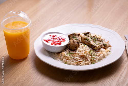 Delicious plate of biryani rice with tender chicken pieces, served with a side of yogurt garnished with pomegranate seeds, and accompanied by a refreshing orange juice in a plastic cup.