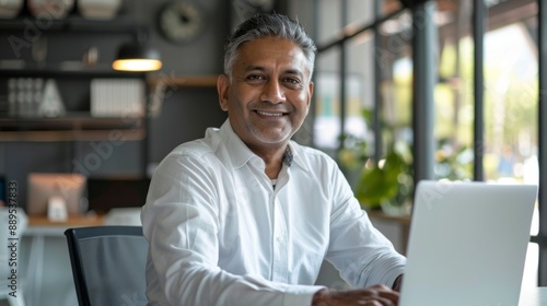 A man in a white shirt is sitting at a desk with a laptop in front of him