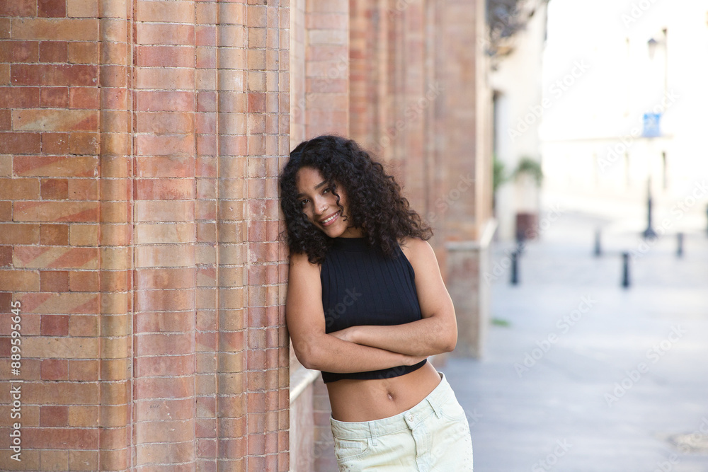Fototapeta premium beautiful Latin woman with black eyes and brunette with long curly hair leaning on her side with her arms crossed on the brick wall. The woman is happy on holiday in Seville, Spain.