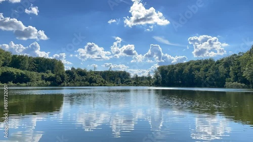 Time-lapse of white clouds moving over peaceful, calm waters of a forest lake. Summer landscape with blue sky and vivid green trees up to the horizon. Concept of calmness, relaxation or mindfulness.