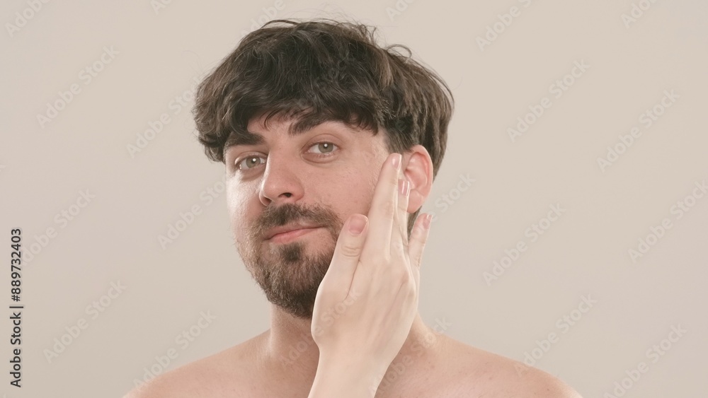 Young adult man looking at camera female applies cream on his face, isolated on white background. Natural beauty skin care concept. Young adult man looking at camera female applies cream on his face, isolated on white background. Natural beauty skin care concept.