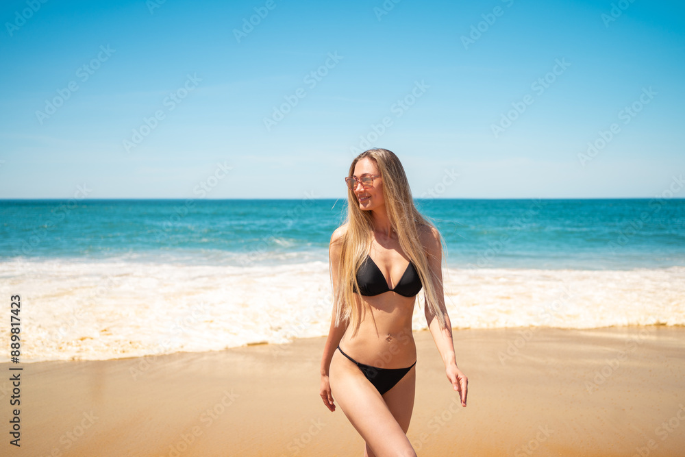 Smiling blonde in bikini walking on sandy beach against sea and blue sky. Attractive young woman in swimwear strolling near shoreline. Lady is enjoying summer vacation on sunny day. Smiling blonde in bikini walking on sandy beach against sea and blue sky. Attractive young woman in swimwear strolling near shoreline. Lady is enjoying summer vacation on sunny day.