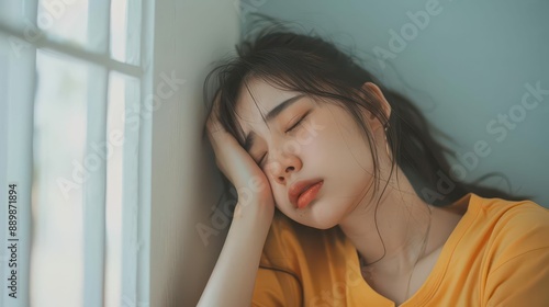 a dizzy asian young woman suffering from a headache leaning against a wall at home expression of discomfort soft indoor lighting healthfocused photo