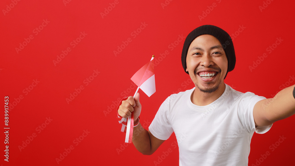 Indonesian youth on white shirt and a beanie hat takes a selfie while celebrating Indonesia Independence Day on August 17 and holding the Indonesian flag, isolated on a red background Indonesian youth on white shirt and a beanie hat takes a selfie while celebrating Indonesia Independence Day on August 17 and holding the Indonesian flag, isolated on a red background