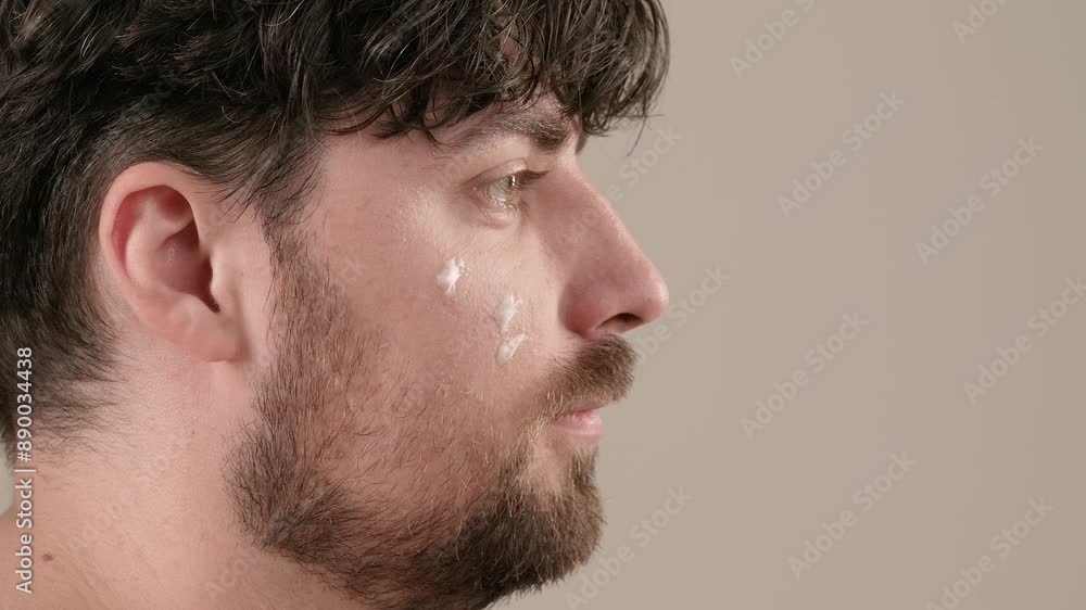 Young adult man looking at camera female applies cream on his face, isolated on white background. Natural beauty skin care concept. Side view. Young adult man looking at camera female applies cream on his face, isolated on white background. Natural beauty skin care concept. Side view.