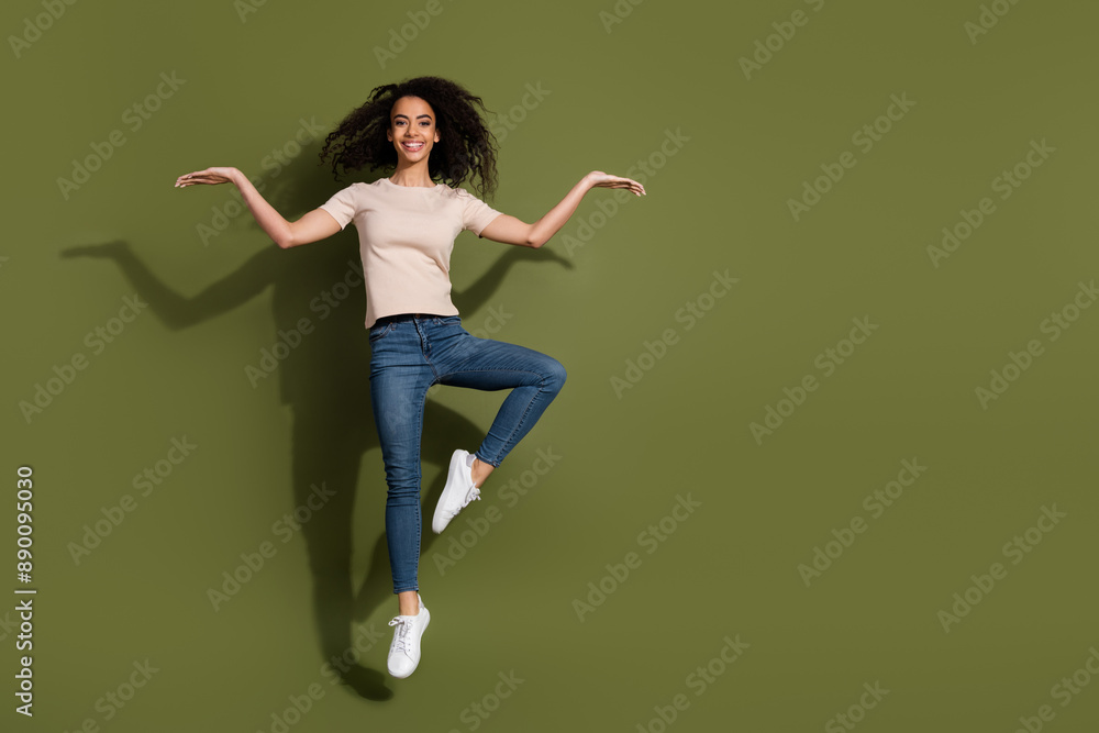 Full size photo of pretty girl dressed beige t-shirt jeans jumping arms presenting objects empty space isolated on khaki color background Full size photo of pretty girl dressed beige t-shirt jeans jumping arms presenting objects empty space isolated on khaki color background