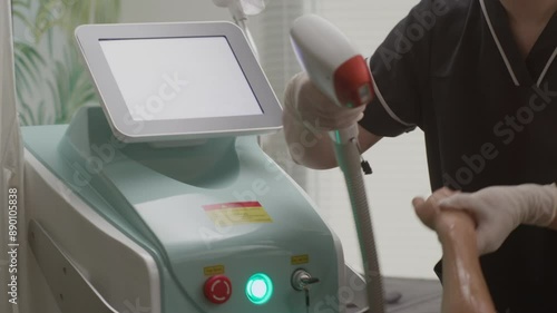 Handheld shot of Asian female specialist setting equipment before procedure of laser epilation on arm of female client at beauty center photo