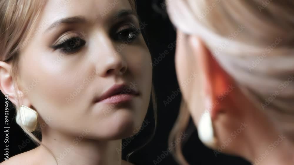 Video of young blonde woman near mirror on black background. Female in black dress with straps and earring. Female beauty makeup concept. Video of young blonde woman near mirror on black background. Female in black dress with straps and earring. Female beauty makeup concept.