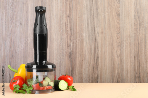 Hand blender kit, fresh vegetables and parsley on beige table against wooden background, space for text