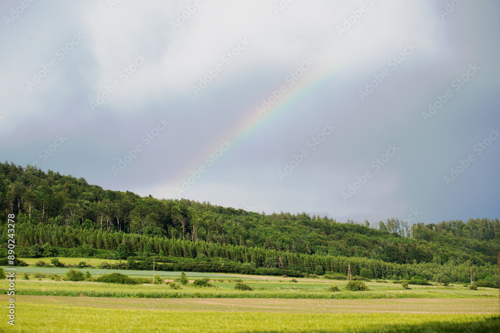 Tęcza nad górami, zielone pola i las na wzgórzu.