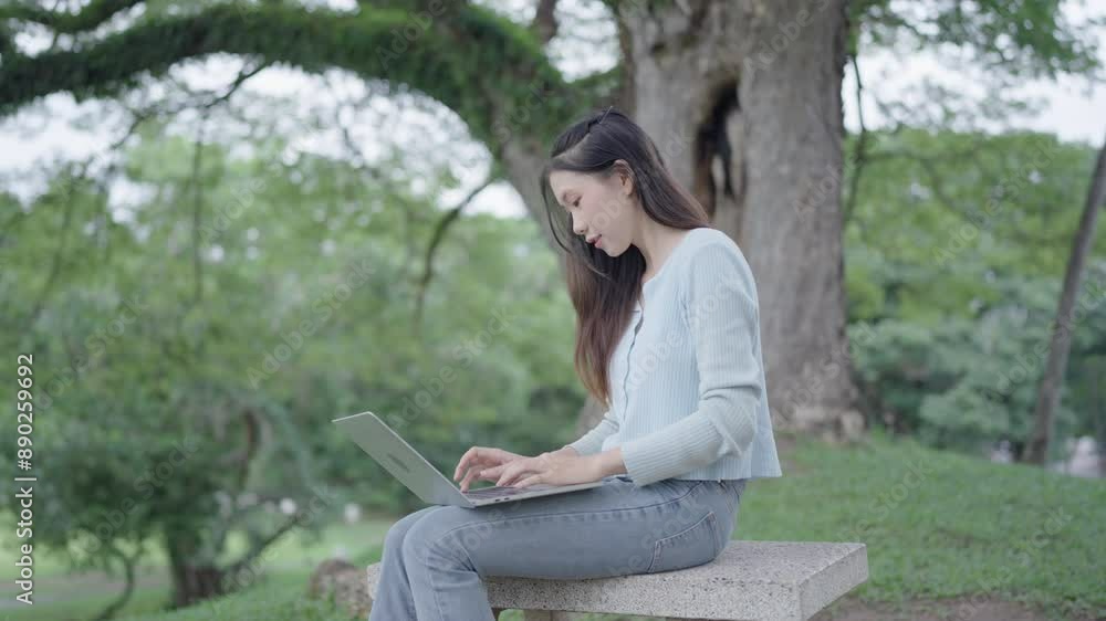 Young focused caucasian woman in casual clothes typing on a laptop while sitting under the tree in a city park Young focused caucasian woman in casual clothes typing on a laptop while sitting under the tree in a city park