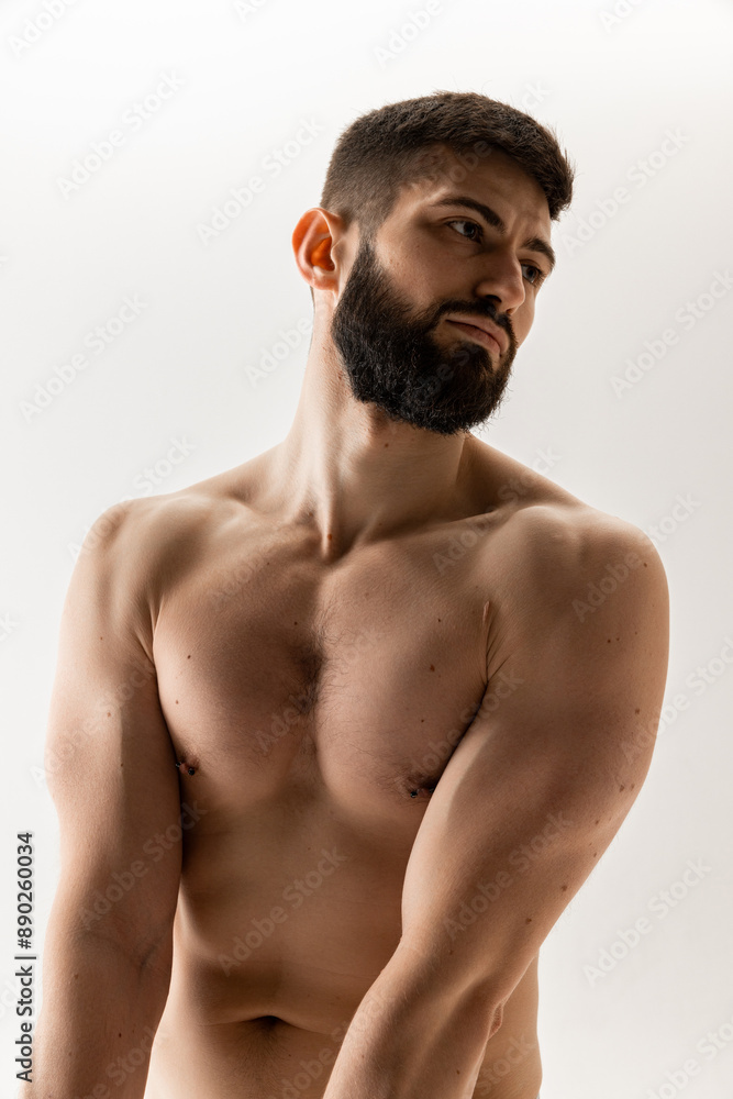 Obraz premium Shirtless young man with beard and short hair looks pensively to side, showing off his muscular physique against white background. Concept of male health, body care, natural beauty people, fitness.
