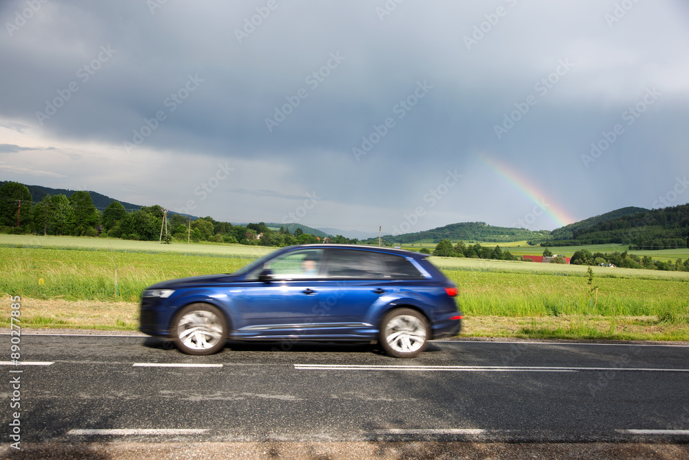 Samochód osobowy na drodze, tęcza nad górami, zielone pola, dachy domów. Samochód osobowy na drodze, tęcza nad górami, zielone pola, dachy domów.