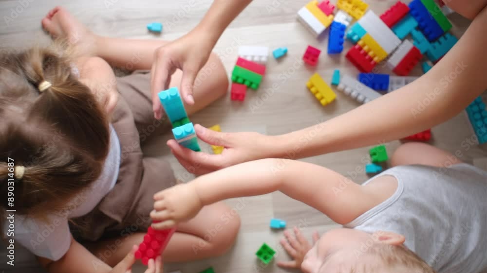 Top view of unknown mother sitting on warm floor with baby son and little daughter teaches children to build tower of colored blocks, family spending free time at home enjoy playtime with toy set Top view of unknown mother sitting on warm floor with baby son and little daughter teaches children to build tower of colored blocks, family spending free time at home enjoy playtime with toy set