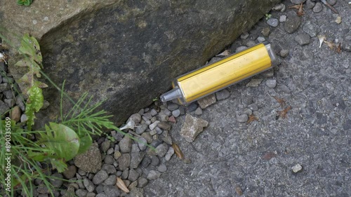 Discarded yellow single-use electronic cigarette vape left on the side of a concrete path with weeds, gravel and rocks.