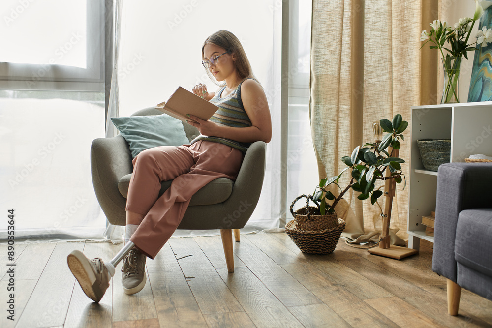 Obraz premium A young woman with a prosthetic leg relaxes in a comfortable chair, engrossed in a book. Natural light floods the modern living room.