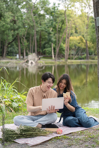 Happy Couple Enjoying a Picnic by the Lake with a Laptop and Snacks in a Serene Park Setting