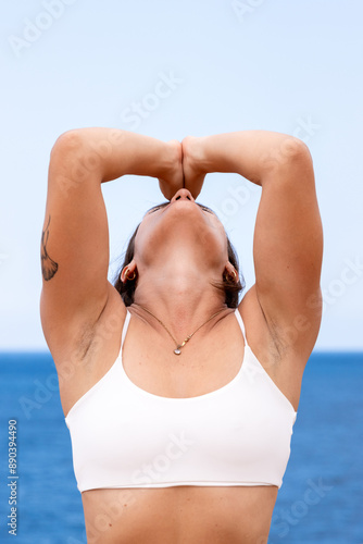 Vertical photo of a girl practicing some yoga exercises in Ibiza with the sea in the background. She has her arms on her face while she is praying to the sky.