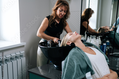 Female hairdresser carefully washing hair of beautiful blonde woman in beauty salon. Beautiful customer lady lying on salon washing bed feeling relax and happy enjoy massaging head.