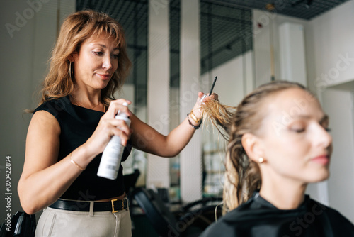 Lady client with long blonde hair getting stylish hairdo done at salon. Female hairdresser spraying hair setting liquid on blonde woman customer hair. Concept of professional fashion hairstyling.