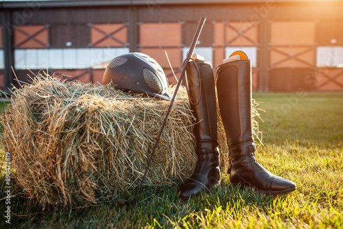 equestrian horse riding equipment with helmet, leather boots and stick on grass and hay bale, barn yard and rancho on background, sunny shot