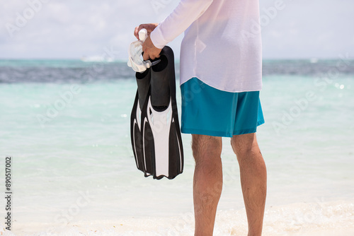 Snorkeling on the reef. Athletically built legs of a man standing on the white sand above the blue sea water and holding swimming flippers in his hand. Fins, vacation. photo